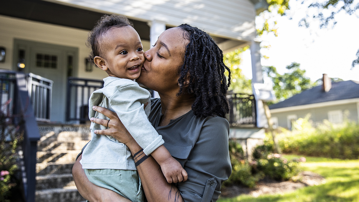 Mother holding child in front of home. 
