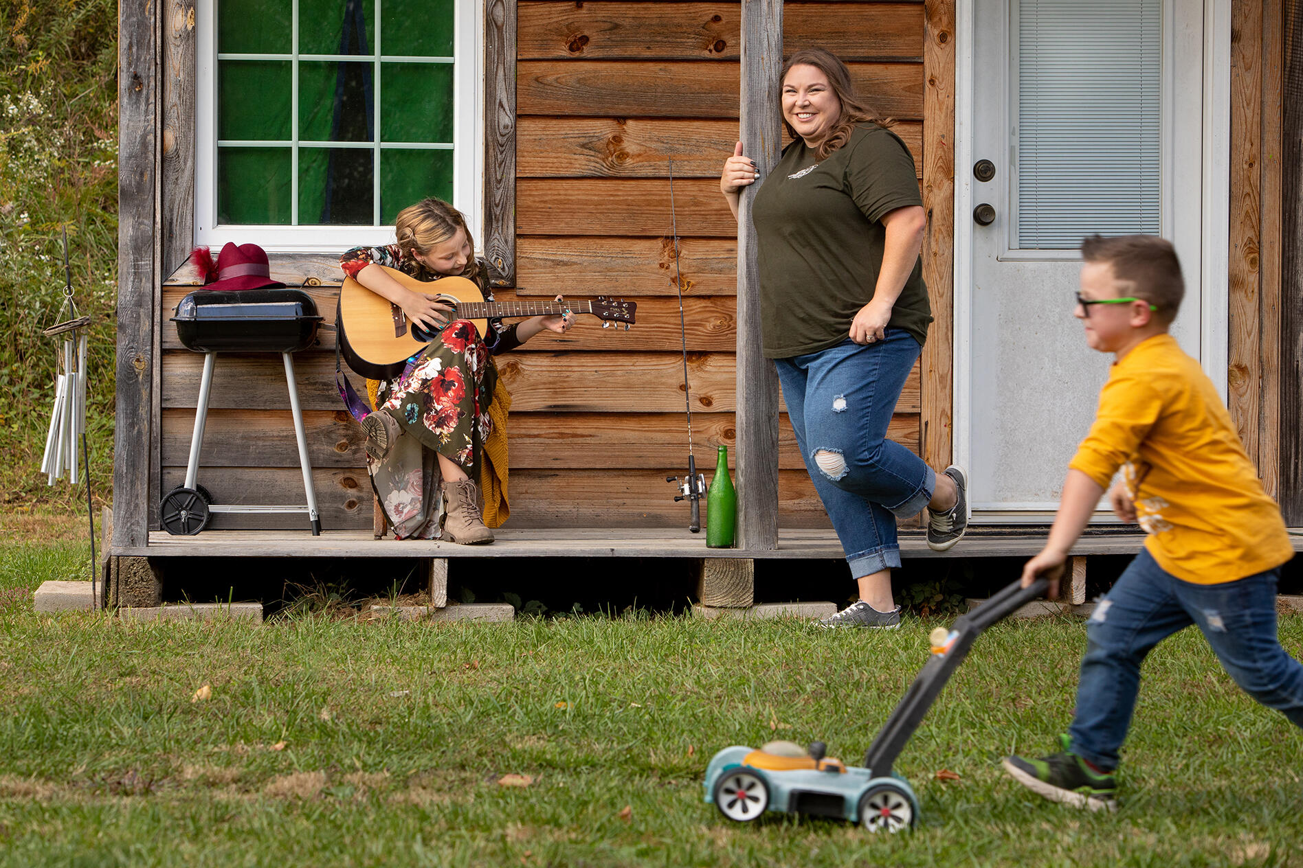 Nine-year-old Alaina Baker plays guitar while her 5-year-old brother, Trevor, pushes a toy lawnmower and their mom, Ashley, watches outside their home. 