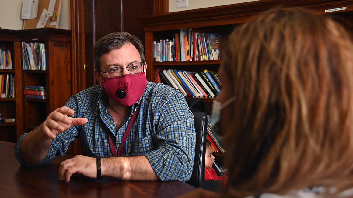 Man in a face mask talking to a women at a table.
