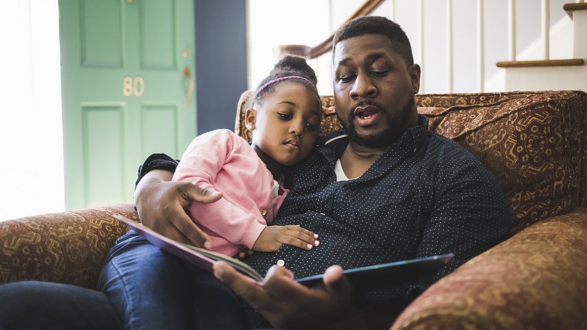 A black father reading to his daughter on their living room sofa. 