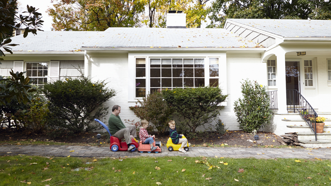 A dad and his two sons riding toy cars in front of their home. 
