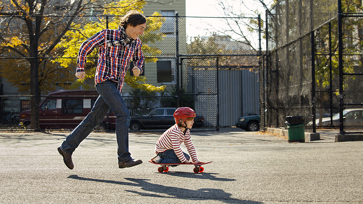 A dad playing outside with his child while they ride a skateboard.