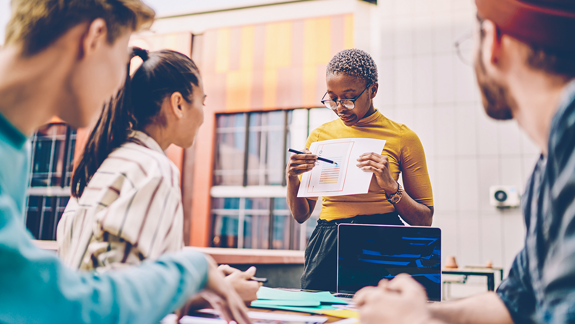 Coach holding paper sketch in hands and explaining data of diagram during workshop for students sitting at one table