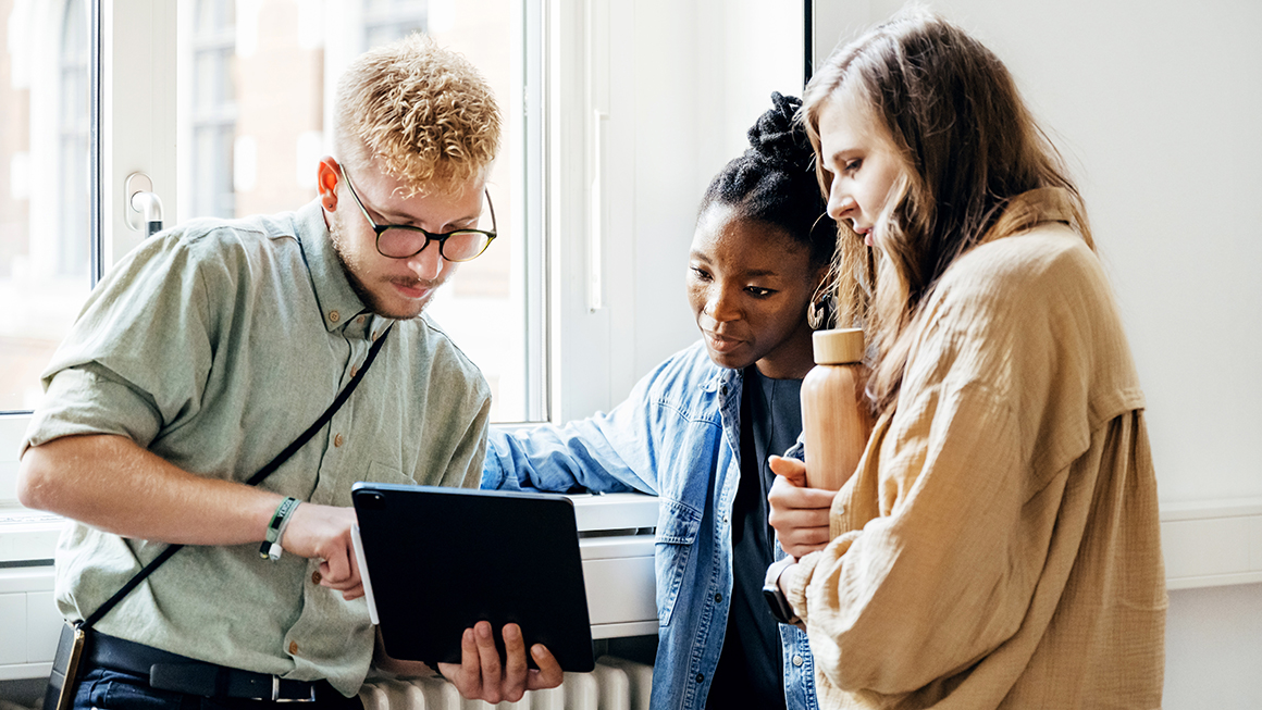 Three young adults huddled around looking at an iPad. 