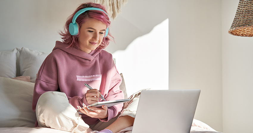 A young girl with pink hair and headphones working on a laptop. 