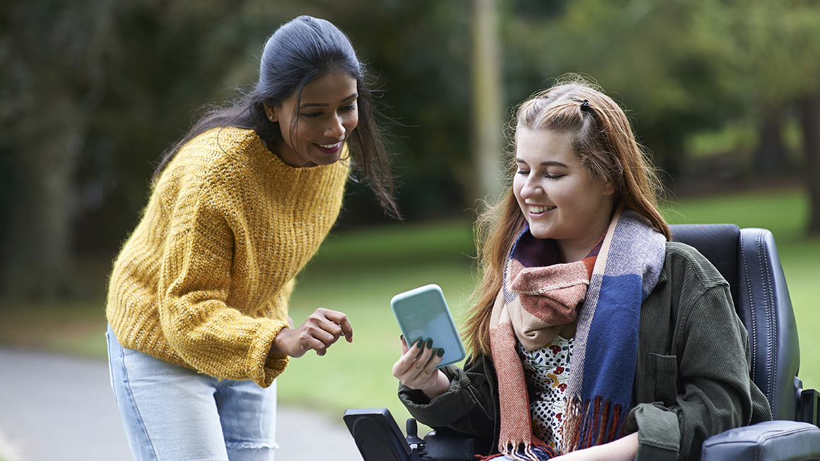 A young woman in a wheelchair talking to another woman and looking at phone together.