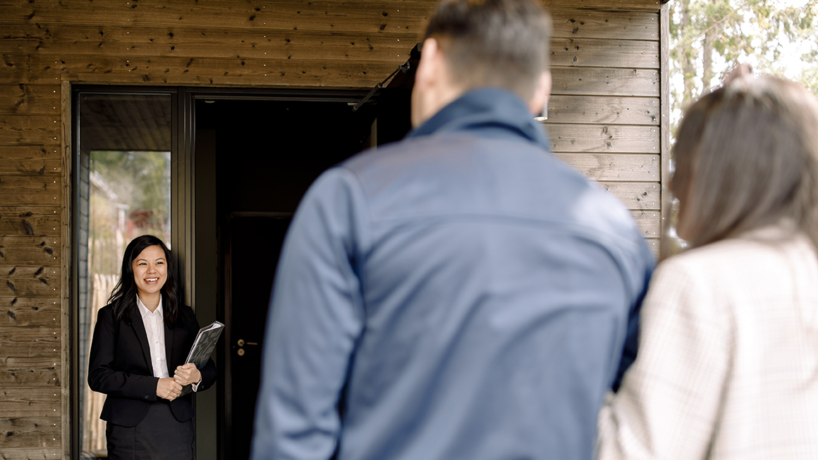 Two people at the front door of an open house and a realtor greeting them.