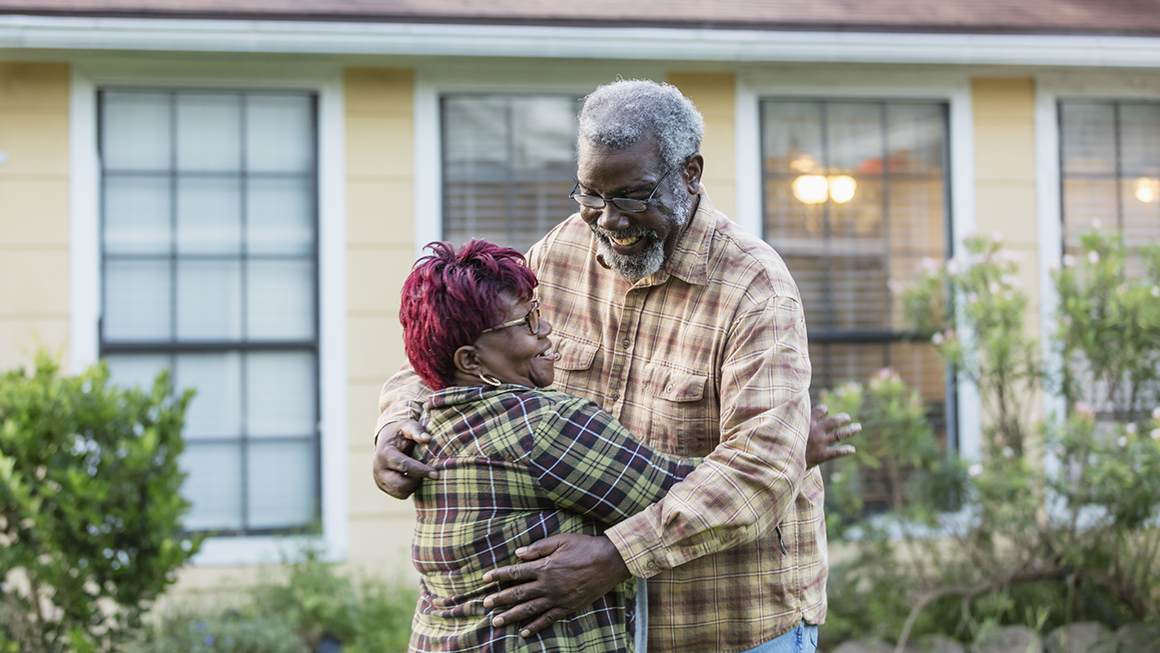 An older black couple hugging in front of housing.