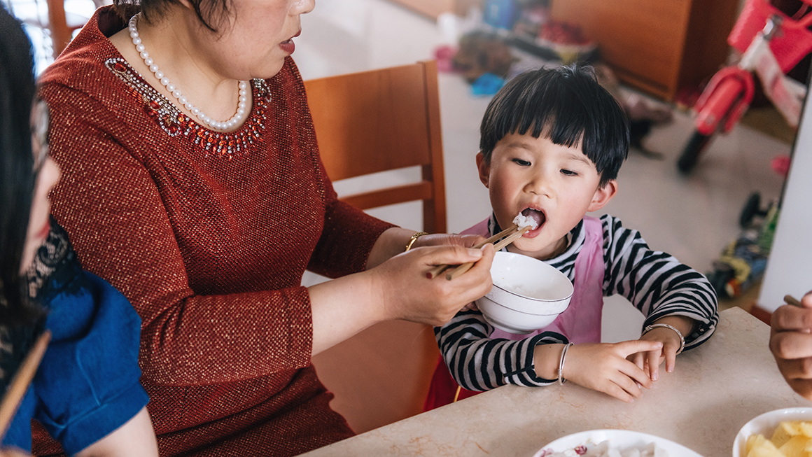 A woman feeding a younger boy some rice.