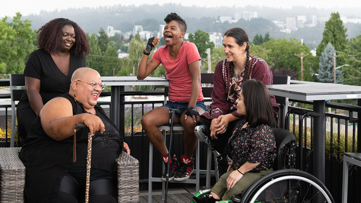 Five disabled people of color with canes, prosthetic legs, and a wheelchair sit on a rooftop deck, laughing and sharing stories. Greenery and city high-rises are visible in the background.