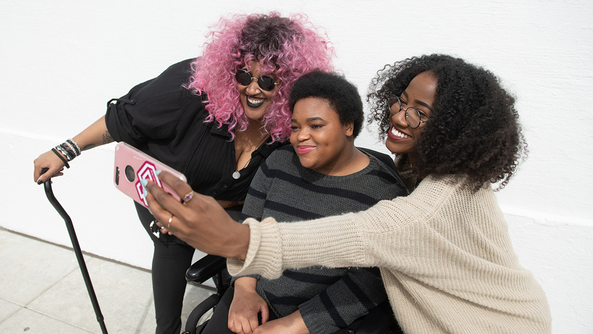 hree Black and disabled friends (a non-binary person with a cane and tangle stim toy, a non-binary person in a power wheelchair, and an invisibly disabled femme) smiling and taking a cell phone selfie together. The photo is angled from the left and cropped around everyone's torso.