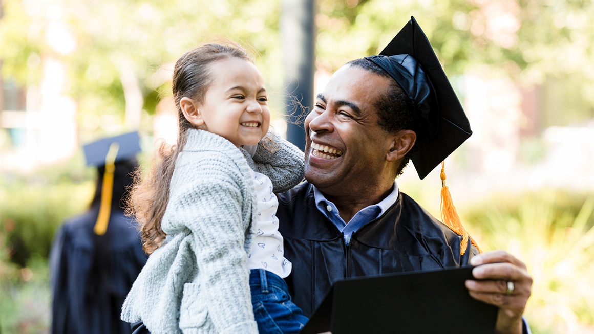 A dad graduating with a cap and gown holding his daughter outside.