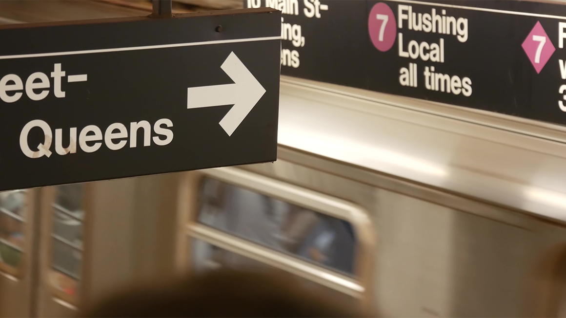 Photo of the inside of a New York subway station, as a subway car is going by. A black sign in the foreground reads "Queens" and points to the platform. Another sign in the distance reads "7: Flushing, Local all times."