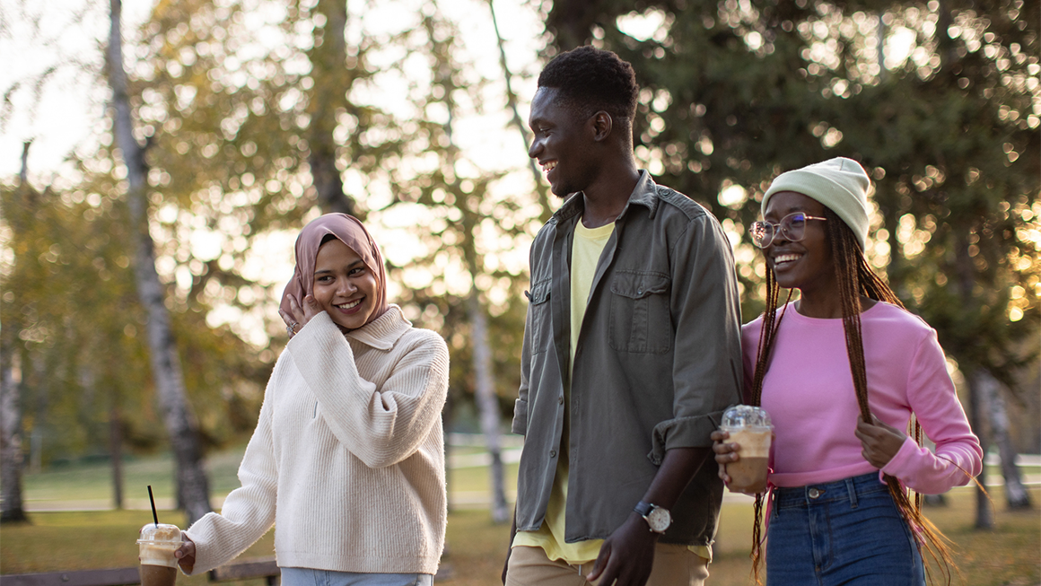 Three friends walk through a park while talking, smiling, and carrying coffee.