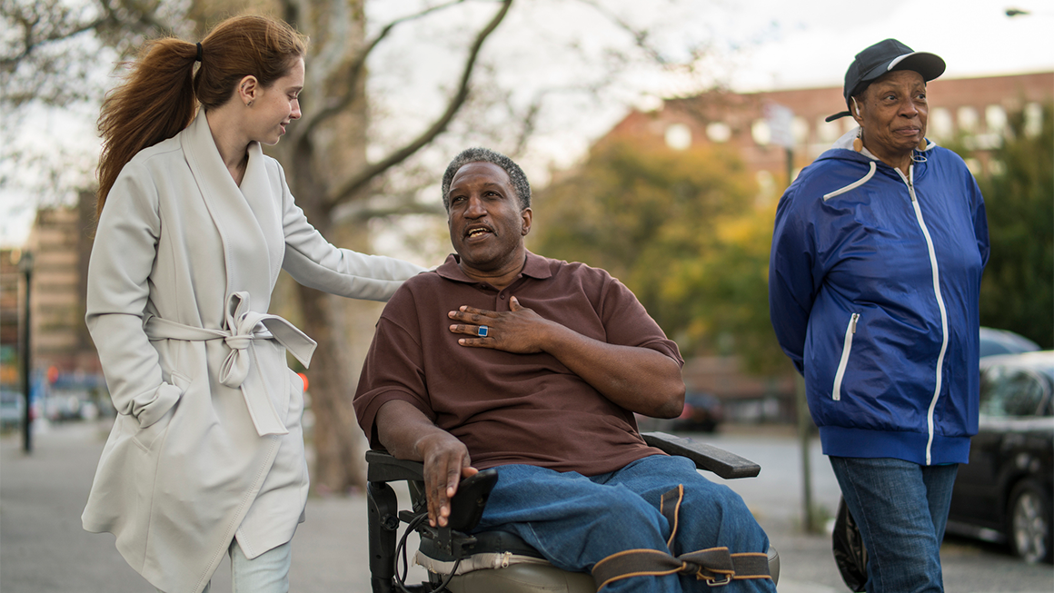 A photo of a young white woman, an older Black man in a wheelchair, and an older Black woman talking as they stroll and roll down the sidewalk together.