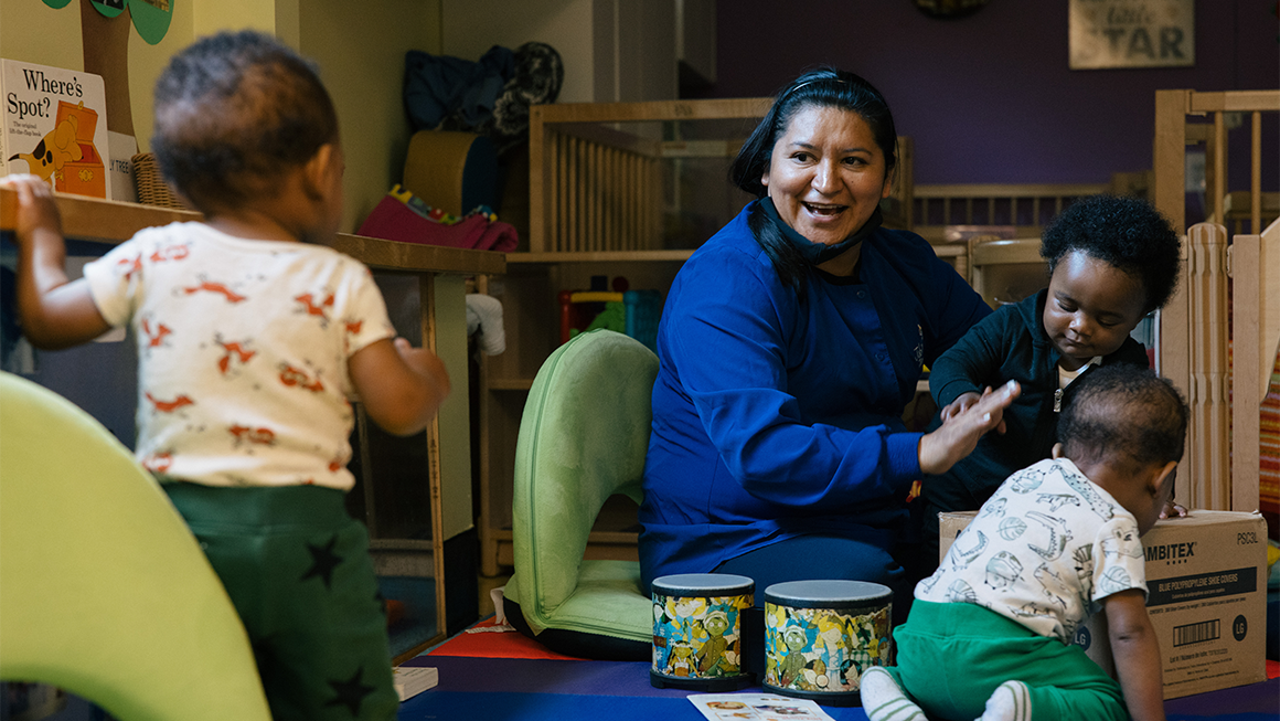 Photo of a child care worker sitting on the ground and smiling as she plays with three toddlers. 