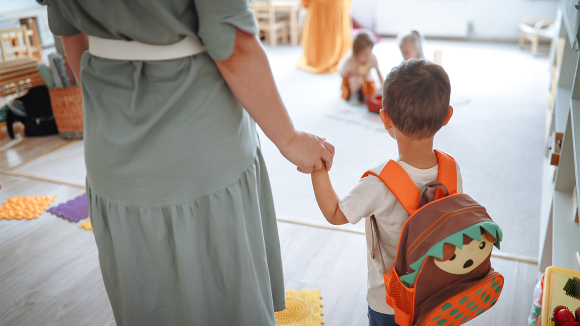 Photo of a toddler wearing a backpack holding his mom's hand as they arrive at daycare.