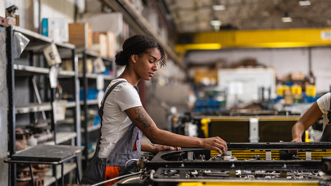 Photo of a young Black woman working on an electric car engine in a factory.