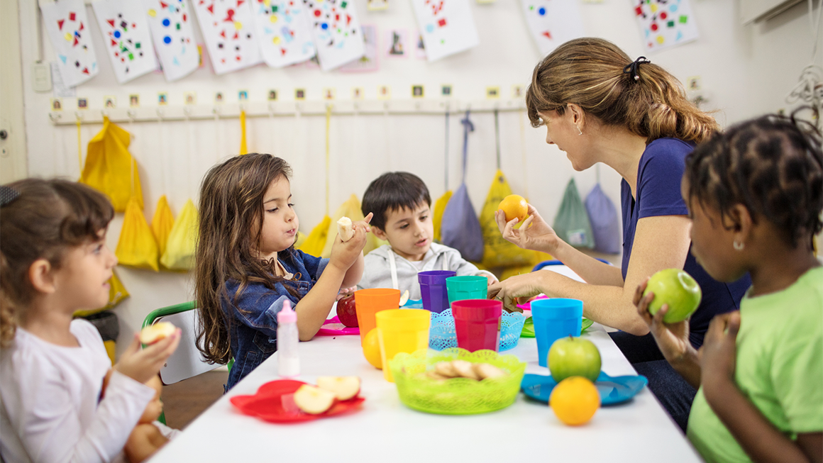 Photo of a young white woman sitting at a table with a diverse group of preschoolers as they eat fruit. 