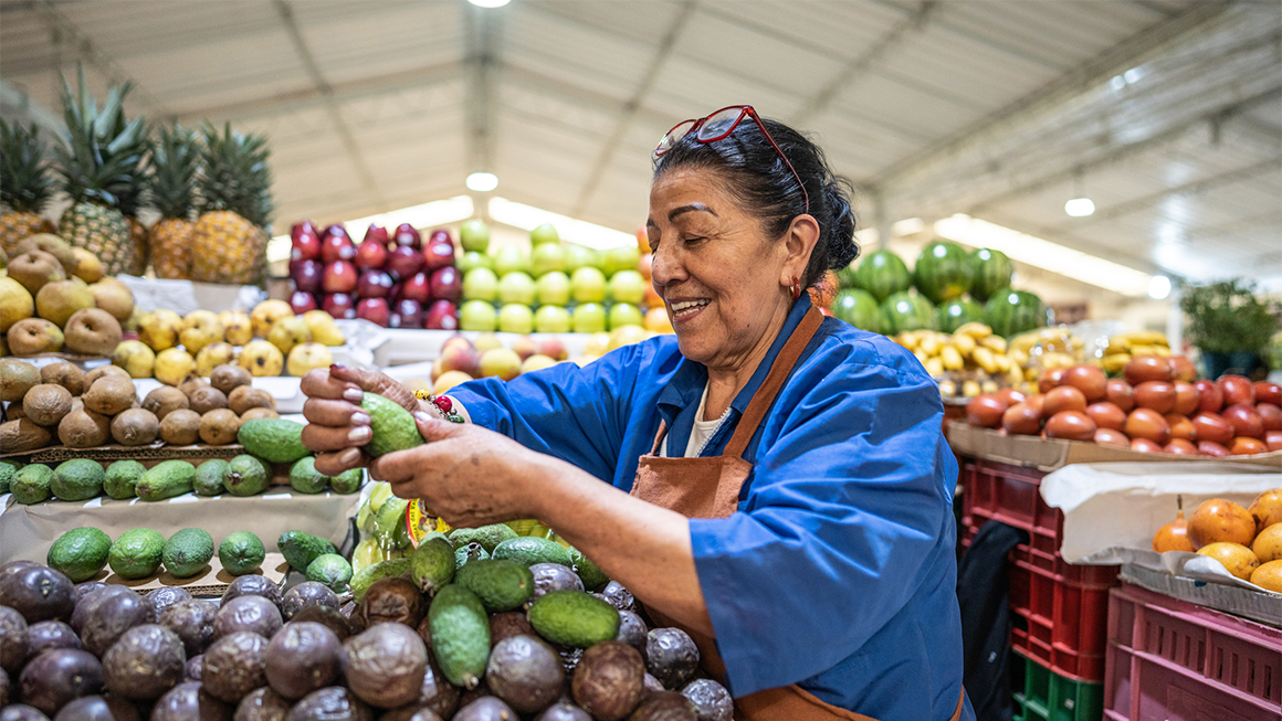 Photo of an older woman restocking avocados at a grocery store.