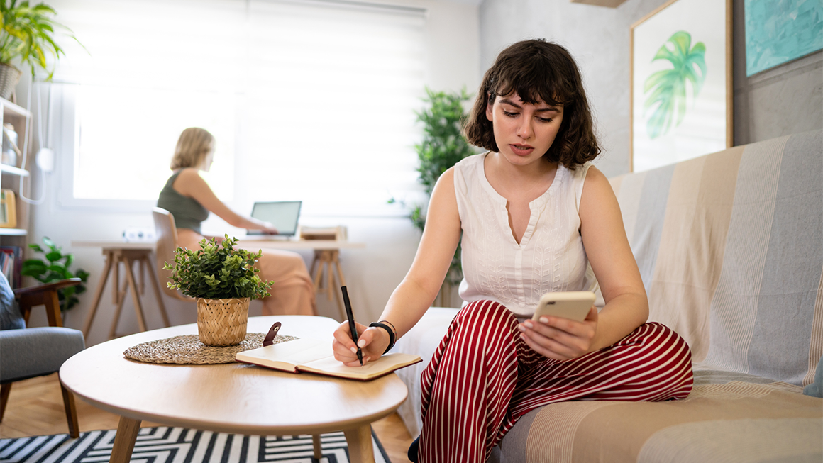 Image of a young white woman checking her phone and writing something down. 