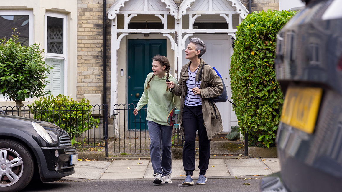 A parent with a backpack and child with a skateboard cross the road on the way to school.