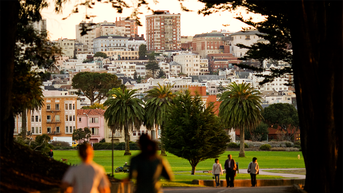 Two people running through a park in San Francisco with apartment buildings and other housing types in the background.