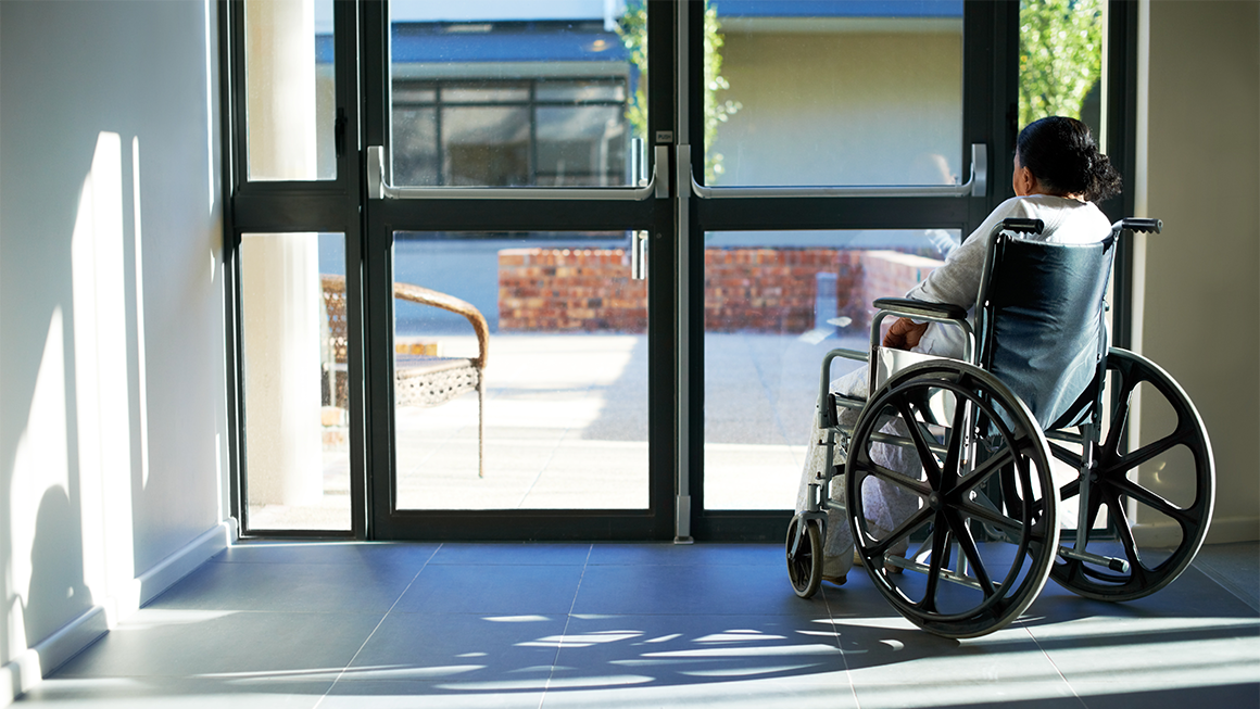 Photo of a woman in a wheelchair looking outside the glass doors of a medical facility. 