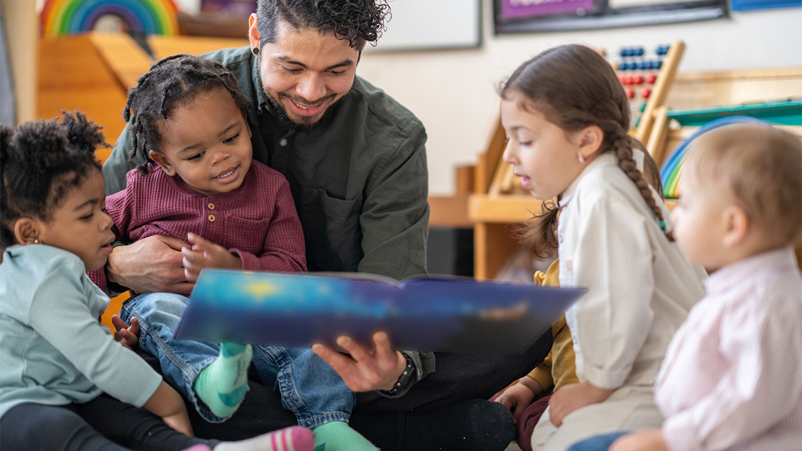 A daycare educator sits on the floor with a child on his lap and other toddlers scattered around him as he reads them a story. They are each dressed casually and are focused on the book.