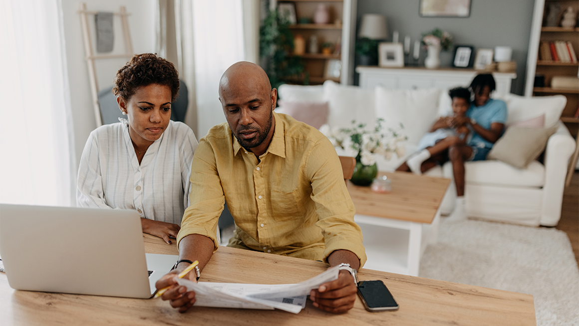 A husband and wife sit at a table and review paperwork together. Their kids sit on a couch playing behind them.