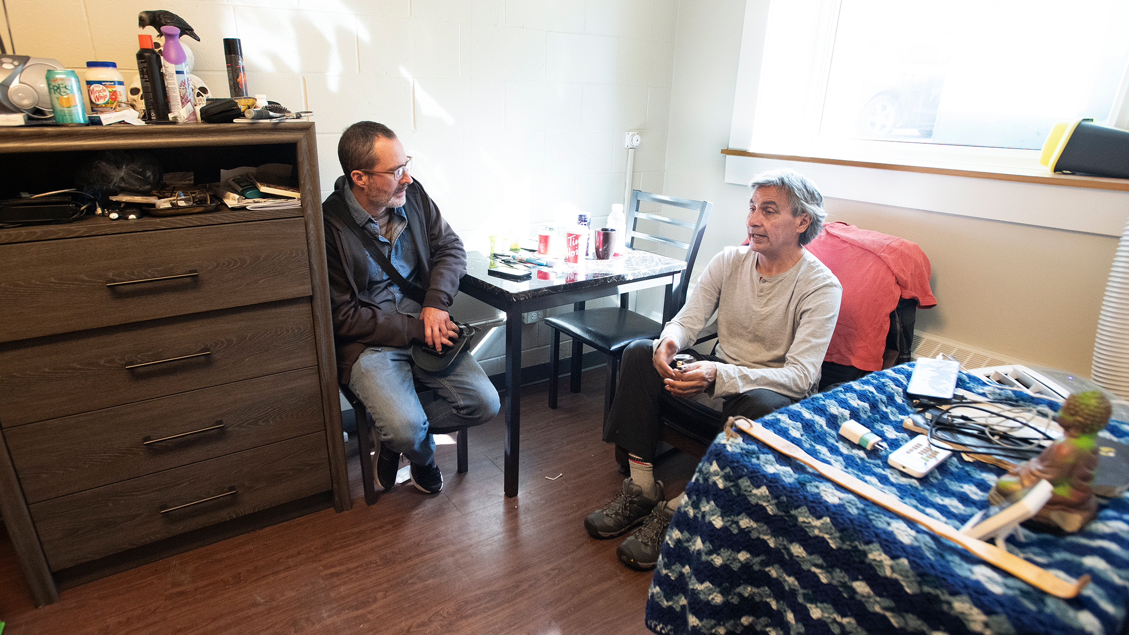 Two men sit in a room at a table beside a bed with personal objects on it