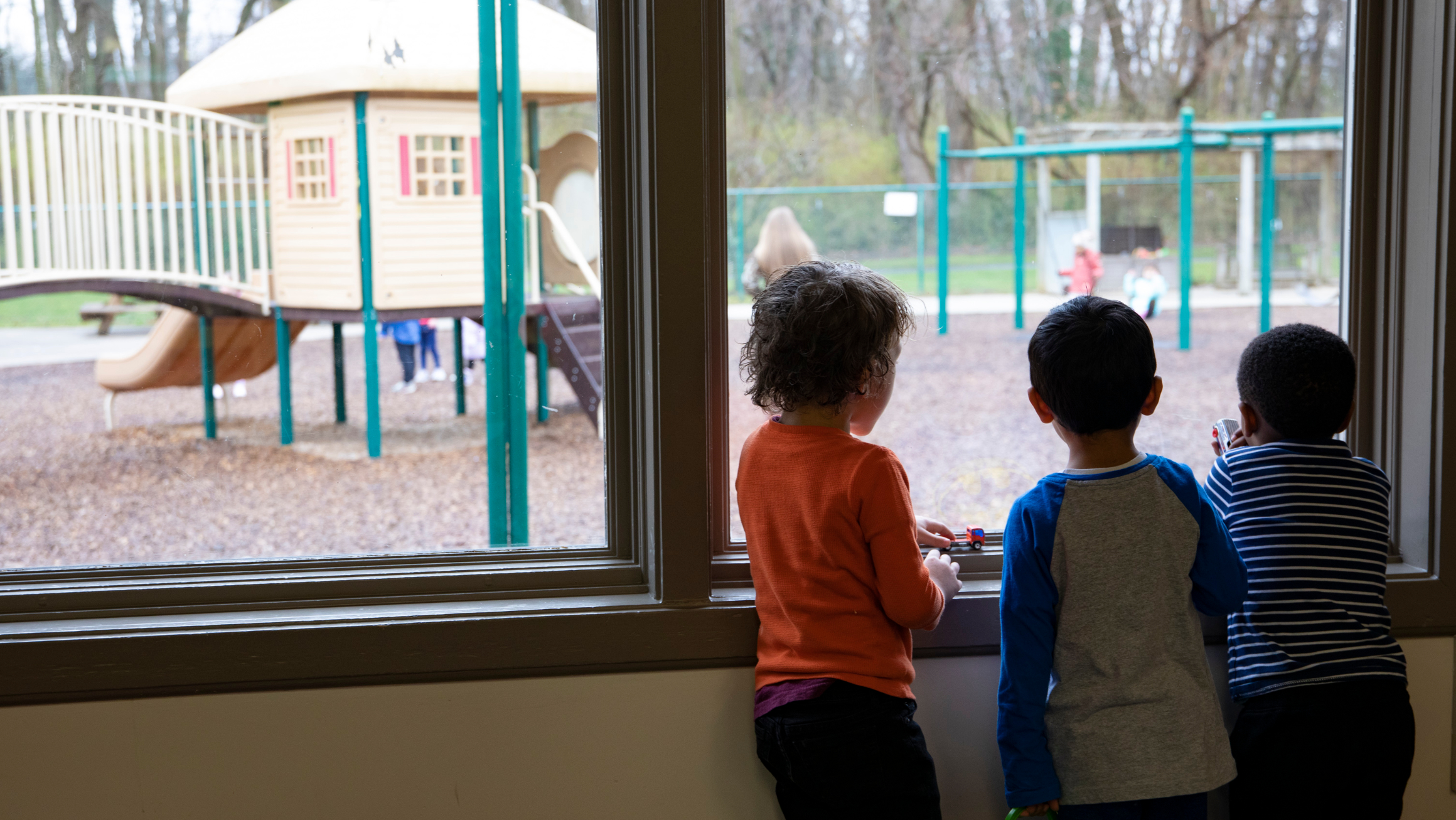 Three children stand at a school window looking outside at a playground