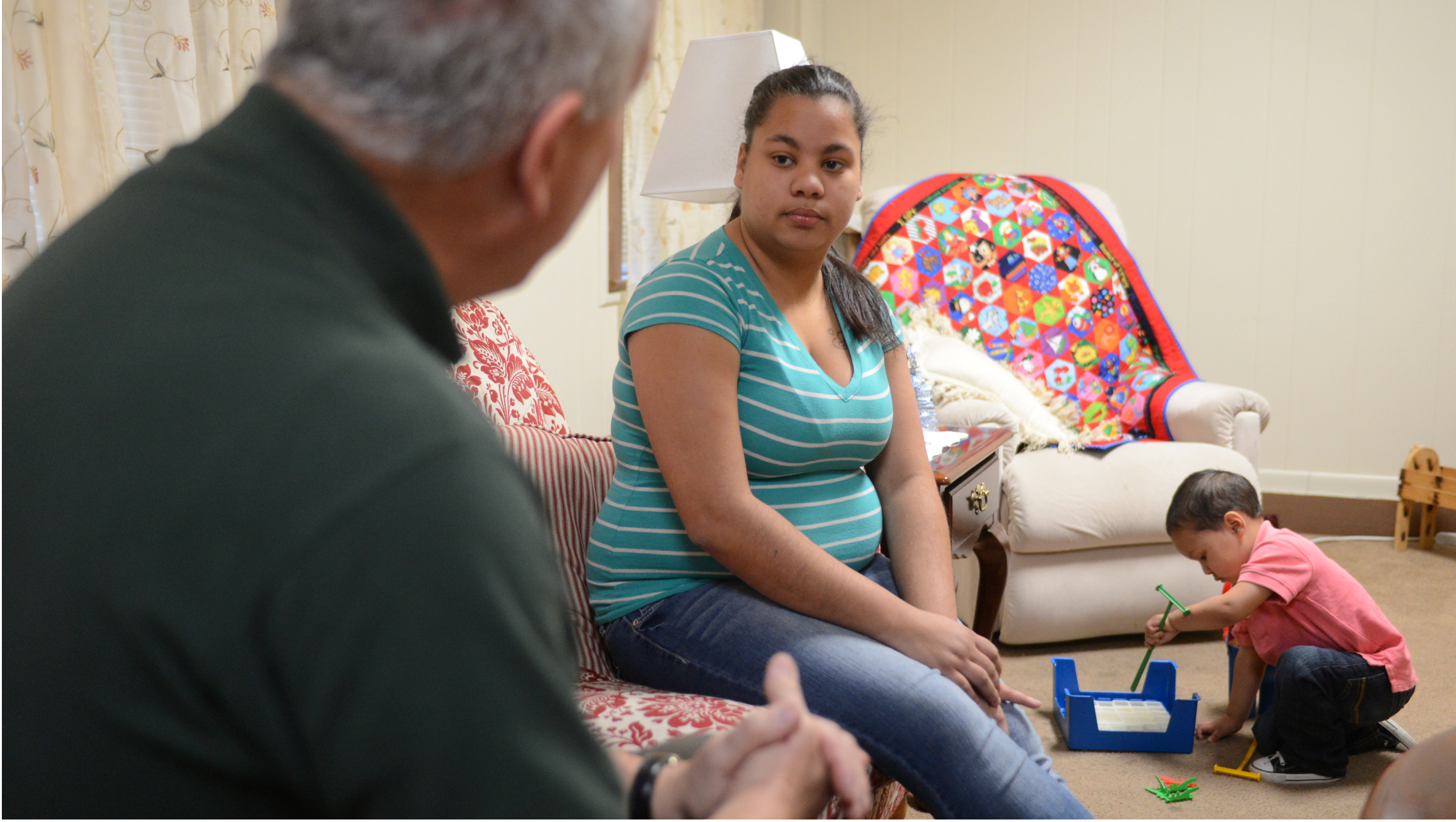 A man and young mother sit on a couch as her toddler son plays on the floor nearby