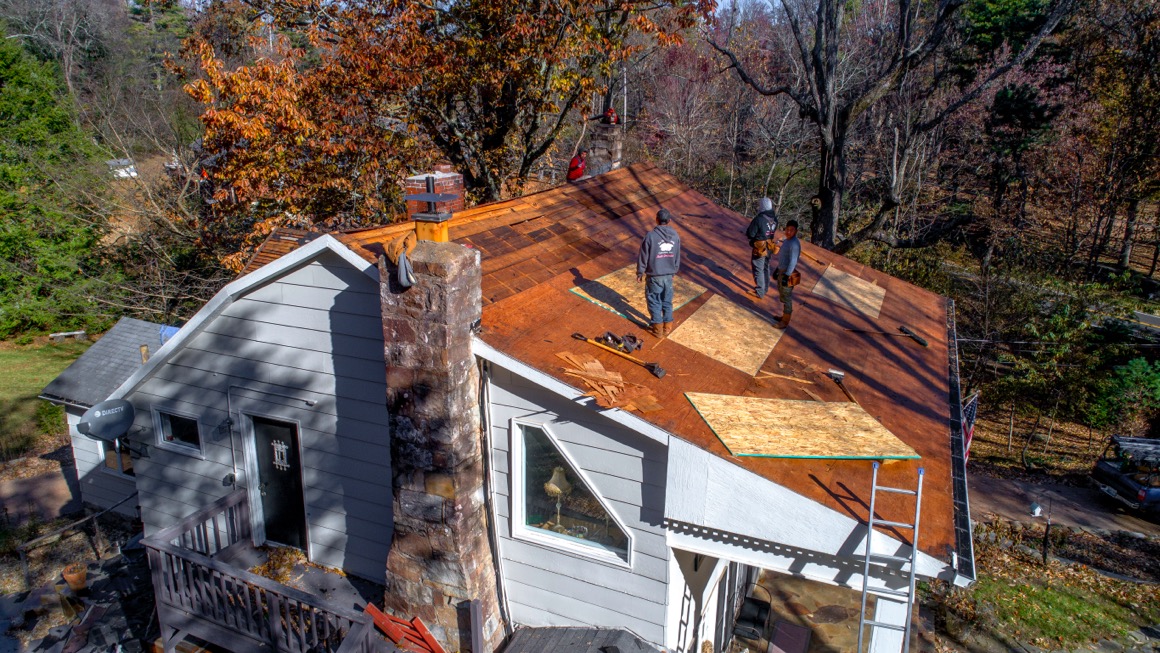 Photo of four construction workers redoing the roof of a small house in a wooded neighborhood.