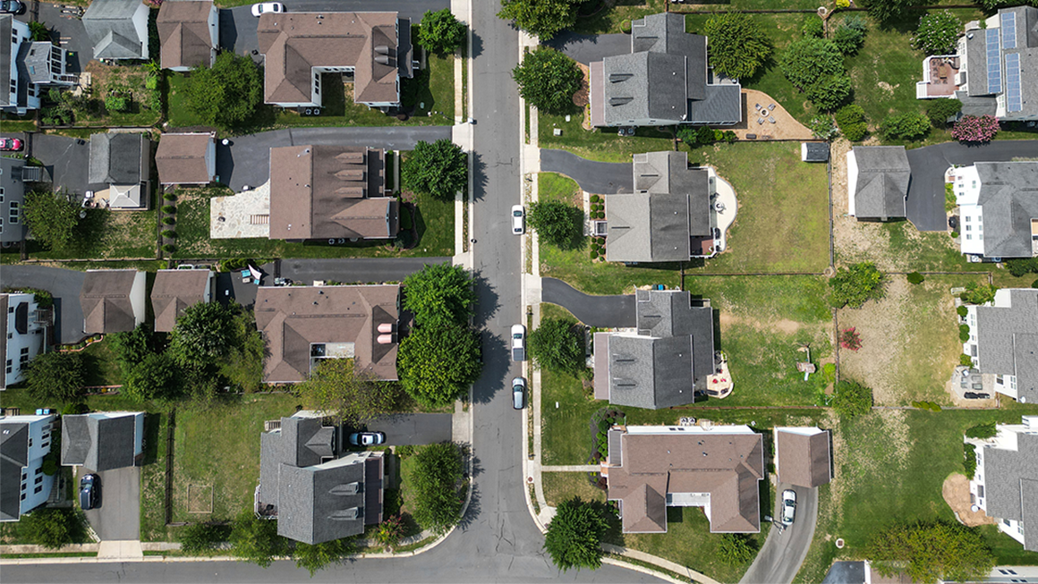 Aerial top-down view of a suburban neighborhood with single-family houses, green lawns, driveways, and trees lining a grid of residential streets, with a few cars parked along the road.