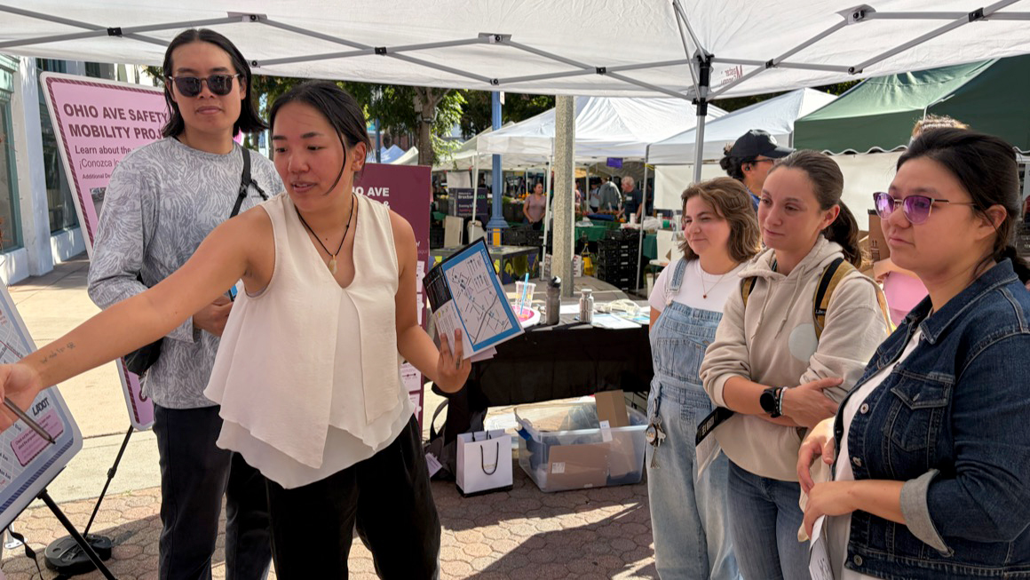 A woman at an outdoor community event stands under a white canopy tent, pointing to a display board while holding a clipboard with a map. Several people stand around her listening. Behind them are tables with materials and a pink sign that reads “Ohio Ave Safety Mobility Project.”