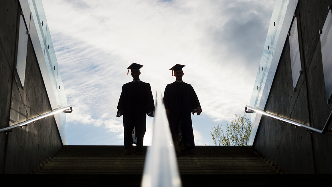 Two graduates in caps and gowns walk down a wide outdoor staircase, silhouetted against a bright sky, with glass railings and concrete walls on either side.