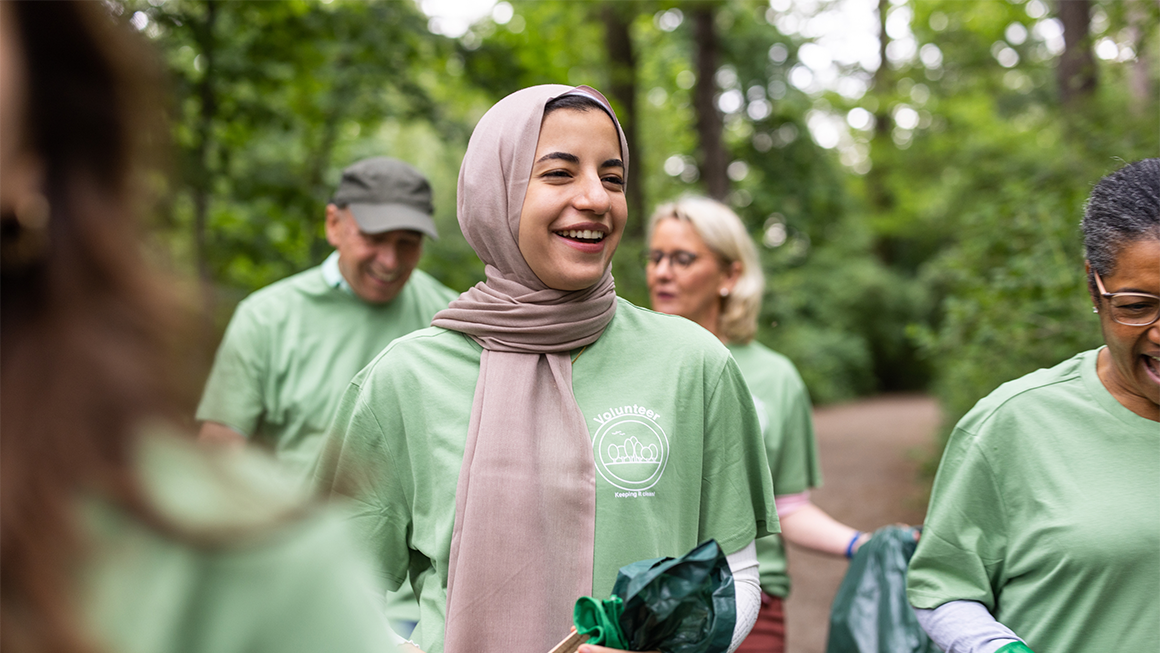 A smiling woman wearing a beige hijab and a green volunteer T-shirt stands outdoors in a wooded area, holding a trash bag, while other volunteers in matching shirts gather around her during a park cleanup.