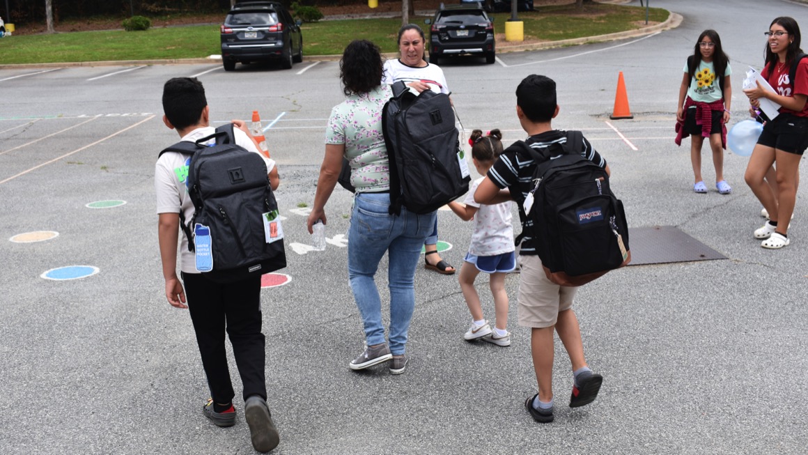 A family of adults and children wearing backpacks walks away in a parking lot 