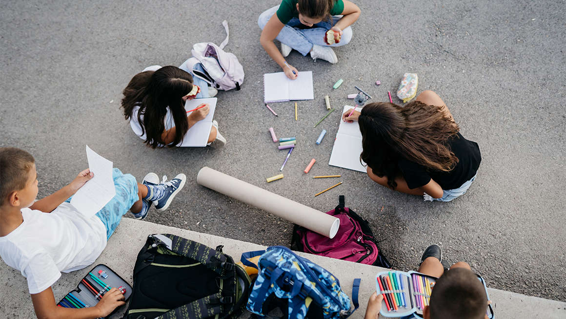 A group of children sit outdoors on pavement in a circle, drawing in notebooks with colorful pencils and chalk scattered around them. Backpacks and art supplies lie nearby, and one child holds a piece of paper while another eats an apple.