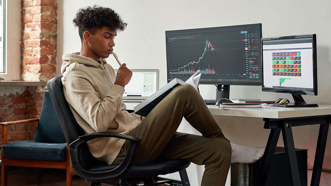 a young man working at a desktop computer at home, reading a book while analyzing financial charts on the screen