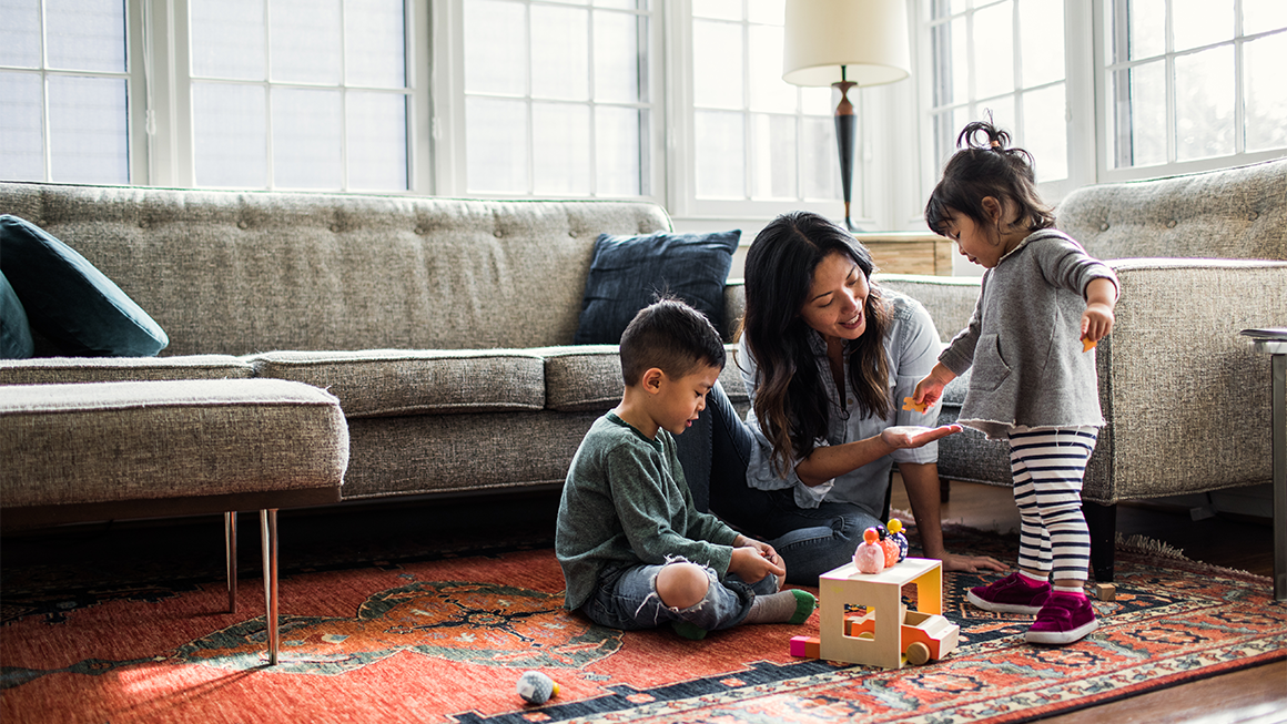 A mother and two toddler children play on the floor of a living room 
