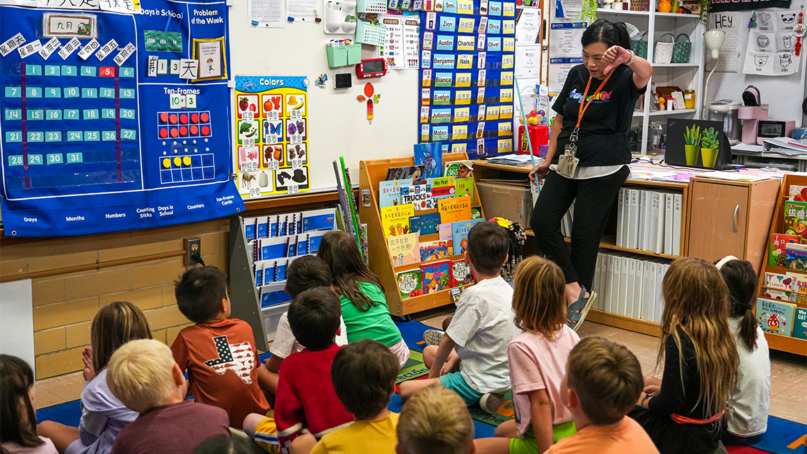 An educator stands at the front of a colorful classroom, reading to a group of young children seated on a carpet, surrounded by learning charts, books, and classroom displays.