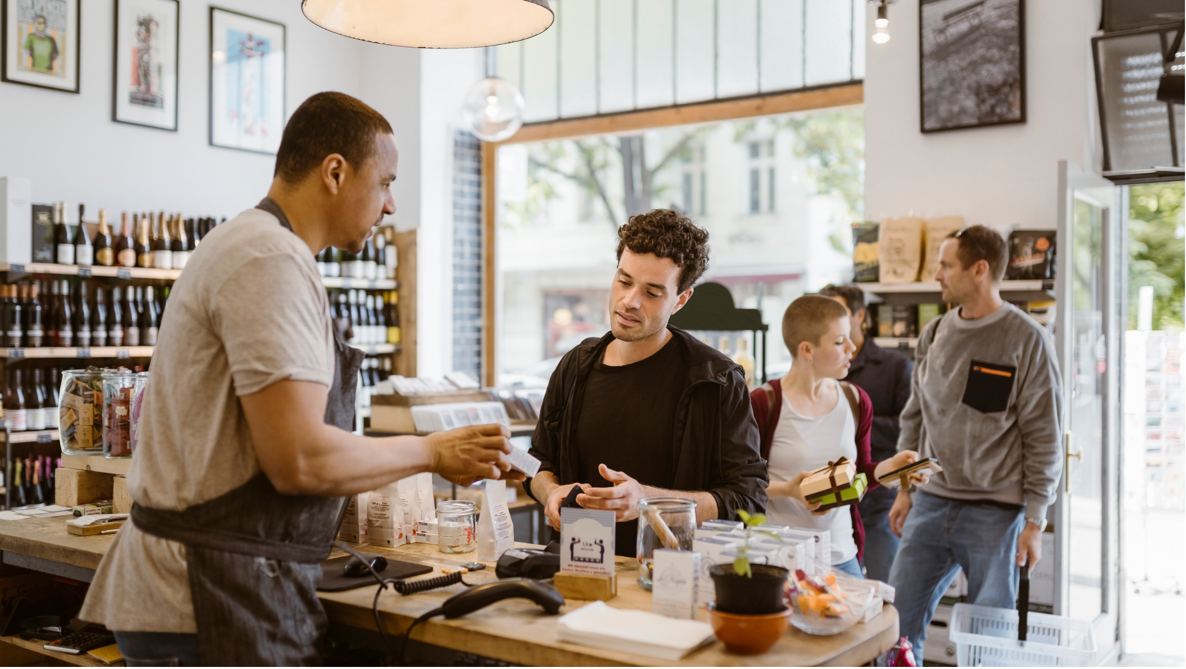 People shopping at a neighborhood store while a cashier assists a customer at the checkout counter.