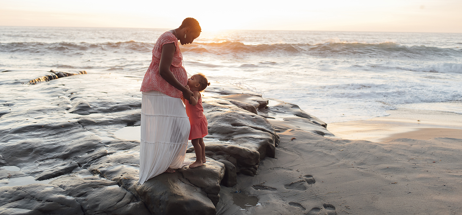 Pregnant person stands with child near ocean