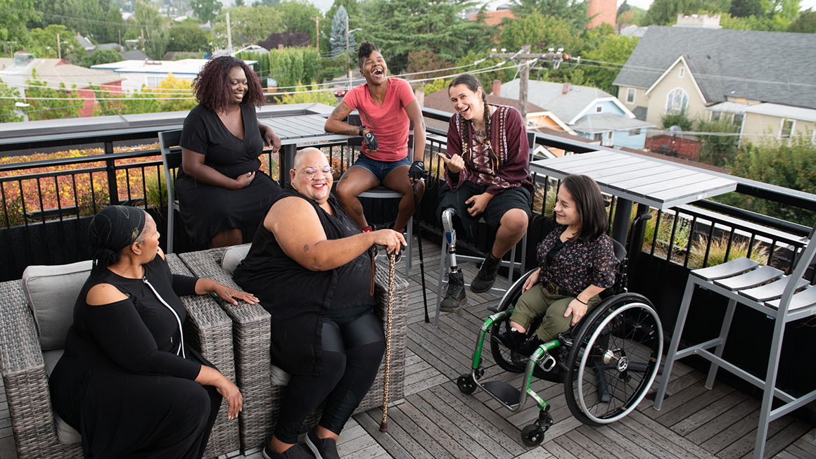 Overhead shot of six disabled people of color at a rooftop deck party. An Indigenous Two-Spirit person with a prosthetic leg smiles directly at the camera and gives a thumbs up while everyone else is engaged in conversation. 