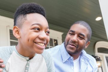Cheerful preteen boy and his dad spend quality time together on their front porch on a sunny day. The father has his arm around his son.