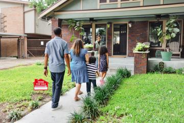 A family of four walks past a "Home for Sale" sign toward a smiling realtor and a red-brick home with an open front door.