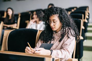A young woman with curly hair, cat-eye glasses, and a light pink hoodie sits among a few other students in a classroom, writing in a notebook with a pencil.