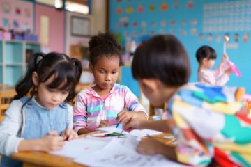 Three young children sit at a table, coloring, painting, and drawing in a colorful classroom.